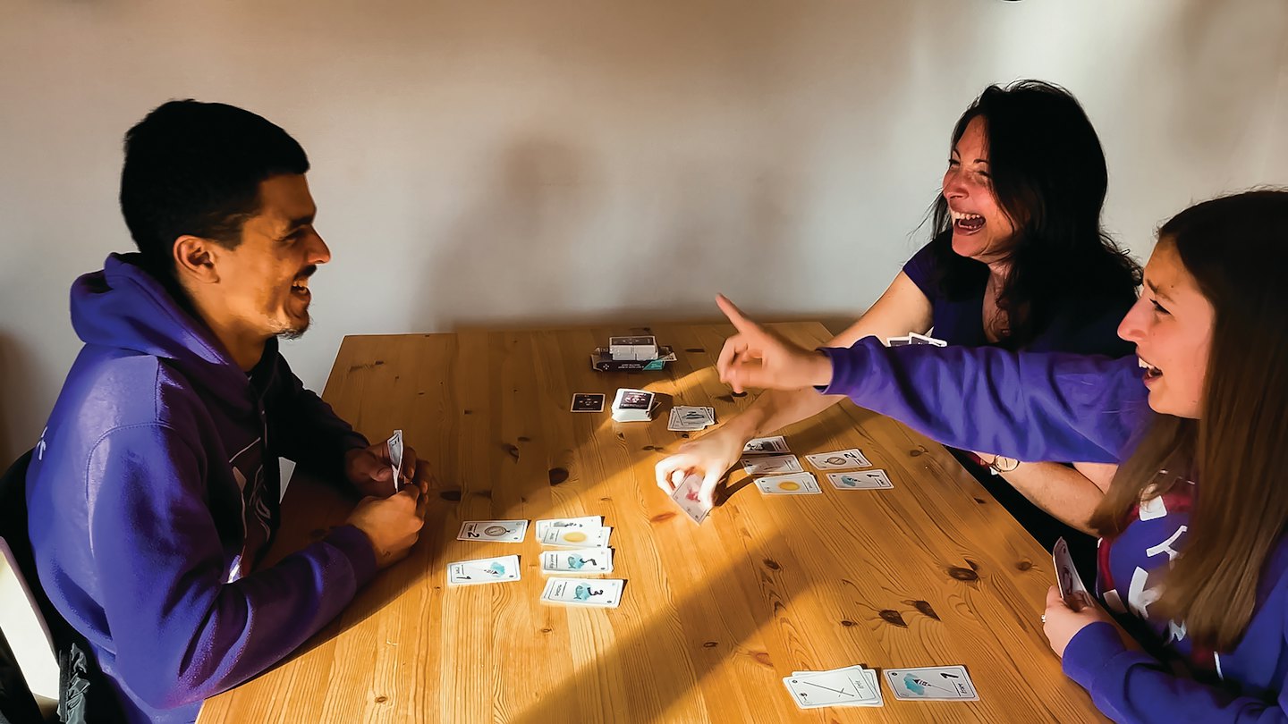 Three friends laugh playing cards at a wooden table.
